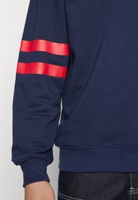 Granatowa bluza z długim rękawem, z dwoma poziomymi czerwonymi paskami na górnej części ramienia, mankiety przy nadgarstkach, noszona z ciemnymi spodniami dżinsowymi.