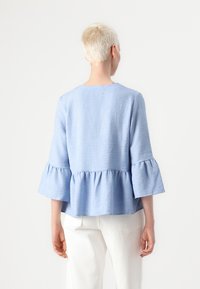 Lyseblå, teksturert topp med trekvart bell-ermer og volangkant, kombinert med hvite bukser, som viser en avslappet passform og enkel design.