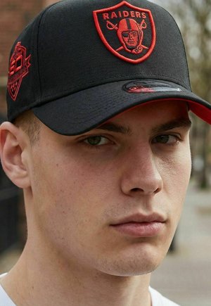 Young man wearing black cap with red Raiders logo and text, looking directly ahead with neutral expression outdoors.
