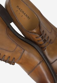 Chaussures habillées en cuir marron avec fermeture à lacets, bout arrondi, détails de couture latérale et semelle marron texturée. Marque Magnanni visible à l'intérieur.