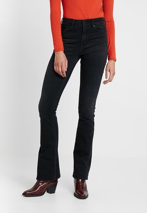 Kobieta w czarnych jeansach bootcut z wysokim stanem, czerwonym topie z długim rękawem i brązowych botkach na obcasie, stojąca na jednolitym białym tle.