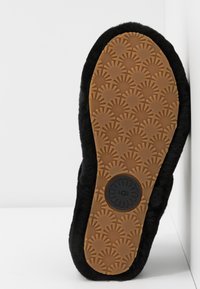 Semelle d'une pantoufle noire avec une semelle en caoutchouc marron à motifs et un tampon de logo circulaire, reposant contre un mur d'angle blanc.