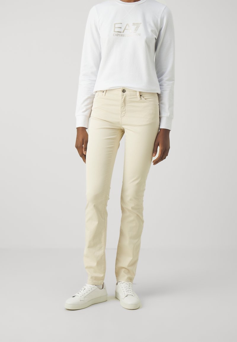 Beiga skinny jeans med framfickor och bälteshällor, tillsammans med en vit sweatshirt med en guldig logotyp. Vita sneakers fulländar stilen.