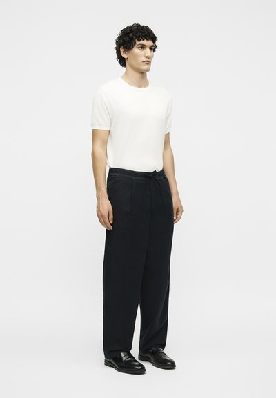 Άνδρας με σγουρά μαλλιά που φοράει απλό λευκό κοντομάνικο πουκάμισο, φαρδιά μαύρη παντελόνια με κορδόνι και μαύρα επίσημα παπούτσια, στέκεται μπροστά σε λευκό φόντο.