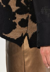 Strickpullover mit einem schwarz-beigen Blumenmuster, kombiniert mit metallisch bronzenen Hosen. Der Pullover hat eine strukturierte Oberfläche und einen gespaltenen Saum.