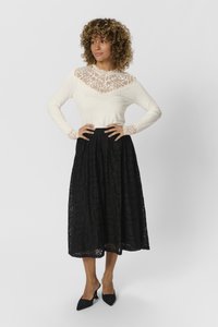 Långärmad elfenbensfärgad topp med spetsdetaljer och en svart spets midi-kjol. Mönstrad tyg med vågiga kanter och blommönster. Svarta klackskor.