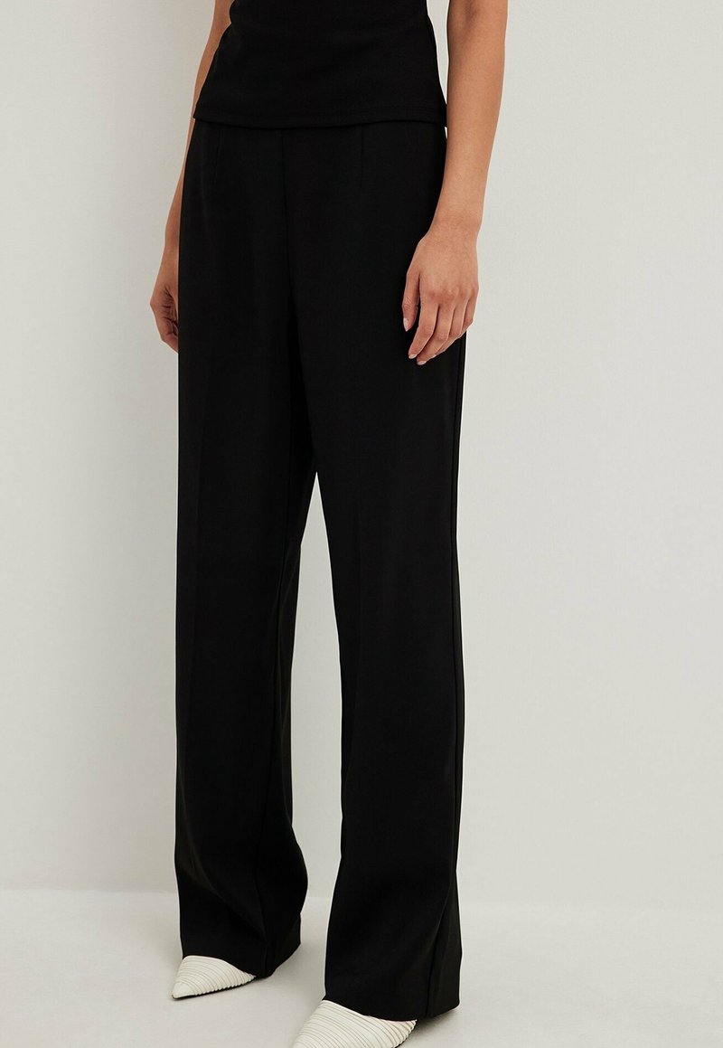 Personne portant un pantalon noir à jambes larges et des chaussures blanches pointues, debout devant un fond clair uni.
