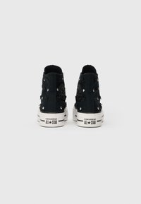 Adidași negri cu botă înaltă de la Converse, cu studuri argintii și fundițe din material, prezentați din spate pe un fundal neutru.