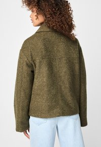 Olijfgroene, textuurgevoelige wolmixjas met een relaxte pasvorm, voorzien van een klassieke kraag en verlaagde schouders. Achternaaddetail zichtbaar.