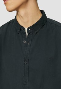 Camisa negra de botones confeccionada en una tela suave y ligera. Presenta un cuello con solapas clásico y un logo bordado "QS" en el pecho.