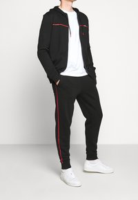 Sudadera negra con acentos rojos y bolsillos con cremallera, combinada con pantalones jogger negros que tienen rayas laterales rojas, usados con zapatillas blancas.