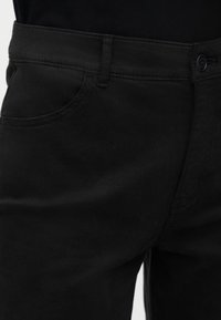 Κοντινή λήψη της μέσης μαύρου τζιν με ορατή μπροστινή τσέπη, θηλιές για ζώνη, κουμπί και φερμουάρ.