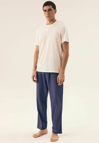 White short-sleeve henley shirt with three buttons and contrasting stitch, paired with loose-fitting dark purple pants, barefoot on plain background.