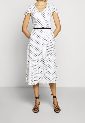 Femme portant une robe midi blanche à pois noirs, manches flottantes, ceinture noire et sandales à talons avec lanières noires, debout les mains derrière le dos.
