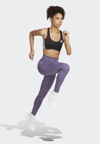 Soutien-gorge de sport noir avec des rayures blanches, leggings violets et chaussures de sport blanches. Modèle dans une pose de course dynamique. Fond clair.