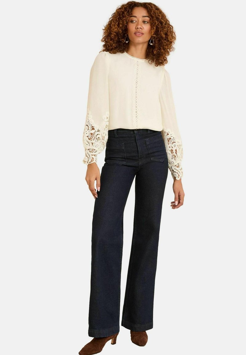 Cream blouse with lace detailing on sleeves, high-waisted dark denim flared pants, and brown heeled shoes. Solid color ensemble.