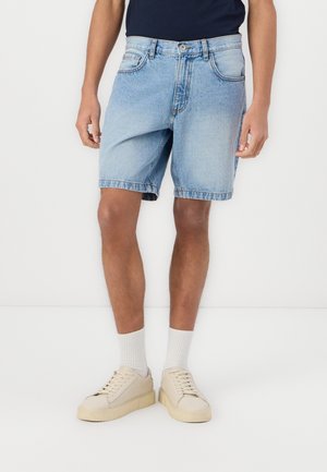 Persoon die lichtblauwe denimshorts, witte sokken, beige sneakers en een donkerblauwe top draagt, staand tegen een witte achtergrond.