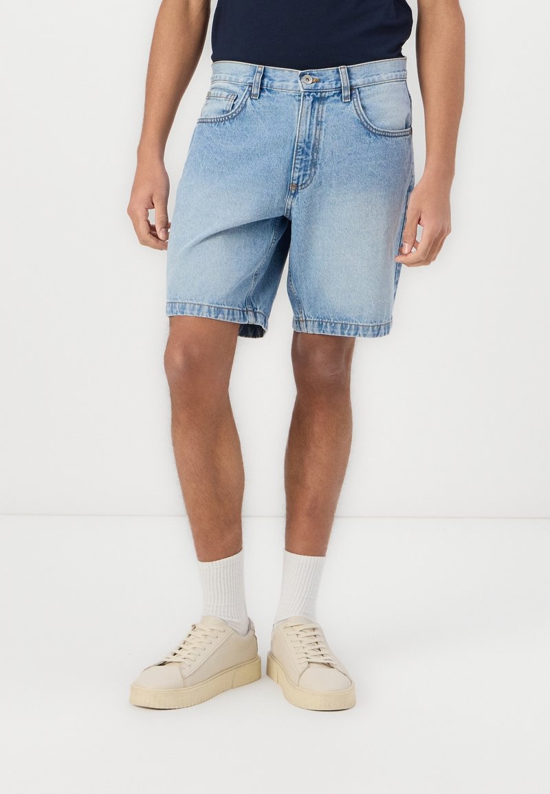 Personne portant un short en jean bleu clair, des chaussettes blanches, des baskets beige et un haut bleu foncé, debout sur un fond blanc.