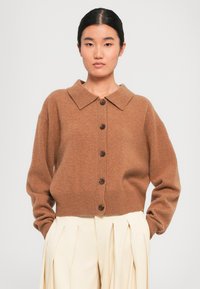 Ung kvinne med mørkt hår iført en brun knappet cardigan med krage og kremfargede, plisserte bukser med høy midje, stående mot en ensfarget bakgrunn.