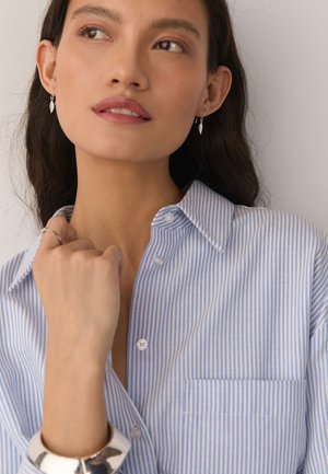 Junge Frau mit silbernen Blatt-Ohrringen, gestreiftem blau-weißem Shirt, einem silbernen Ring und einem breiten silbernen Armband, die zur Seite blickt.