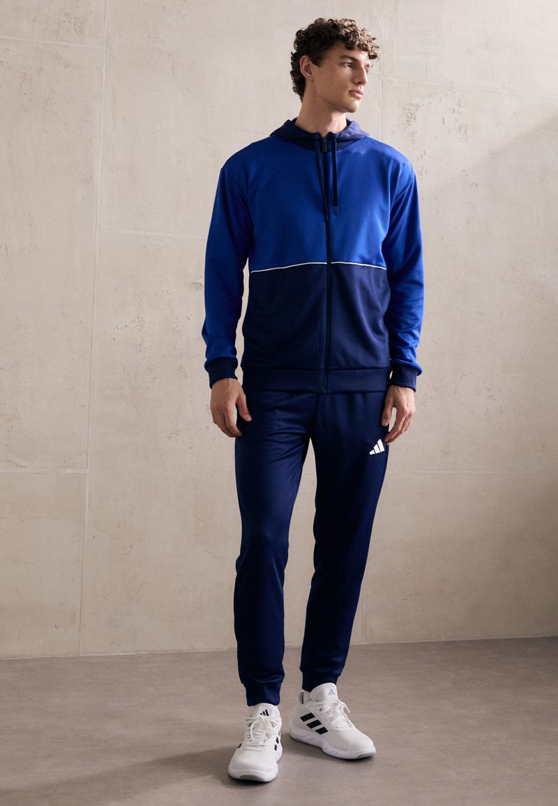 Plavi i tamnoplavi trenerka s duksericom na zip i hlačama s elastičnom trakom u struku, s bijelim naglasnim crtama i Adidas logom na hlačama.