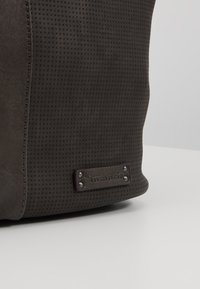 Coin d'un sac marron foncé texturé avec une pièce en cuir cousue et des clous en métal embossés du logo "Tamaris" sur une surface neutre.