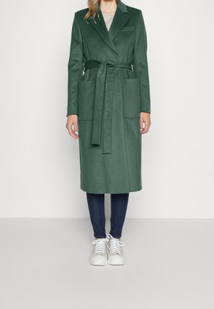 Abrigo largo verde oscuro con cinturón y grandes bolsillos delanteros, usado sobre vaqueros oscuros y zapatillas blancas en una persona de pie.