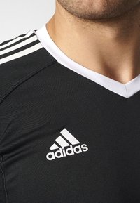 Maillot de sport noir avec des finitions blanches, présentant un design en V, trois rayures blanches sur l'épaule et un logo Adidas brodé en blanc.