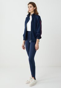 Giacca a zip blu navy con colletto alto, maniche con polsini, leggings abbinati e sneakers bianche. Presenta una striscia a contrasto sulle maniche.