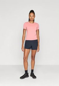 Kvinne som står iført en rosa kortermet skjorte, svarte shorts, svarte sokker og svarte joggesko foran en ensfarget lys bakgrunn.