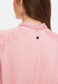 Rosafarbene Bluse aus strukturiertem Stoff mit einem Rüschensaum am Kragen, kleinem Punktmuster und einer Knopfdetaillierung am hinteren Halsauschnitt.