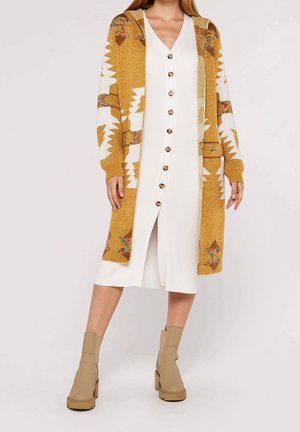 Vrouw die een lange witte jurk met knopen draagt, een mosterdgele gebreide cardigan met capuchon en beige enkellaarzen, staand tegen een effen achtergrond.