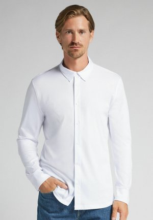 Man met lichtbruin haar draagt een wit shirt met lange mouwen en blauwe spijkerbroek, poserend met een hand in zijn zak tegen een lichtgrijze achtergrond.
