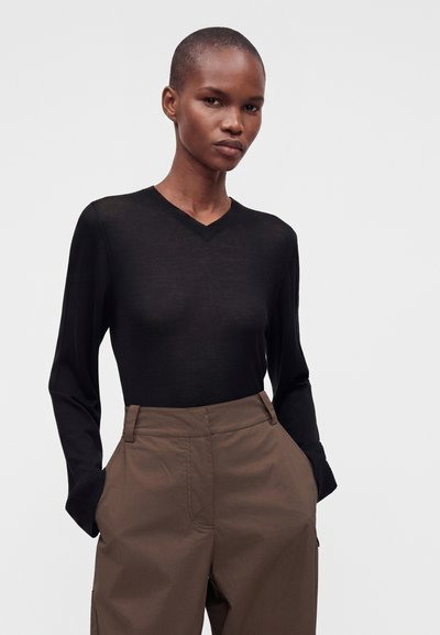 Femme aux cheveux courts portant un haut noir à manches longues et un pantalon marron taille haute, les mains dans les poches, debout devant un fond clair.