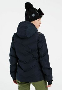 Marineblauwe puffer jacket met capuchon, voorzien van een gewatteerd ontwerp, elastische manchetten en een strakke, moderne snit. Gekleed met een gebreide zwarte beanie.