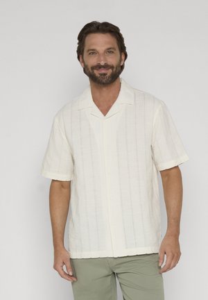 Mand med skæg iført en off-white kortærmet skjorte med knapper og olivengrønne bukser, stående mod en ensfarvet baggrund.