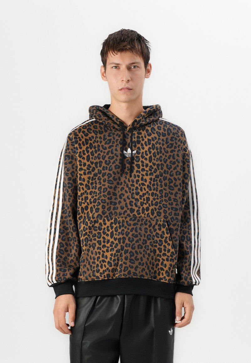 Giovane uomo in posizione frontale che indossa una felpa con stampa leopardata marrone e nera e righe bianche sulle maniche, insieme a pantaloni neri con logo.