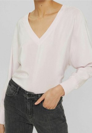 Person trägt eine hellrosa langärmlige V-Ausschnitt-Bluse, die in eine hoch taillierte schwarze Jeans gesteckt ist, mit einer Hand in der Tasche.