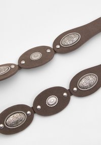 Brauner Wildlederriemen mit ovalen Metallelementen, die filigrane Designs und silberfarbene Beschläge aufweisen, verbunden durch kreisförmige Perlendetails.