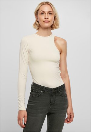 Beige dlouhý rukáv bodysuit s asymetrickým střihem na jednom rameni, spárovaný s černými džíny s vysokým pasem. Hladká textura látky.