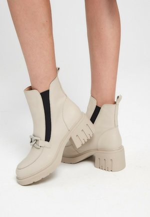Beige Stiefeletten mit klobigen Absätzen, schwarzen elastischen Seitenteilen und dekorativer Schlaufenverzierung, getragen an nackten Beinen vor einem schlichten weißen Hintergrund.