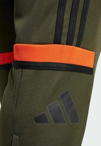 Zelene atletske hlače s crnim trakama, narančastim naglascima i istaknutim crnim Adidas logom. Teksturirana tkanina s prianjajućim dizajnom.