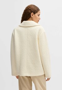 Texturerad krämfärgad jacka med krage, lös passform och synlig sömdetalj; ihop med ljusa byxor.