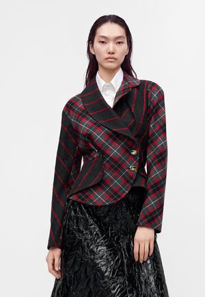 Femme portant une veste rouge et noire à carreaux avec des boutons dorés sur une chemise blanche et une jupe noire texturée, debout devant un fond blanc.
