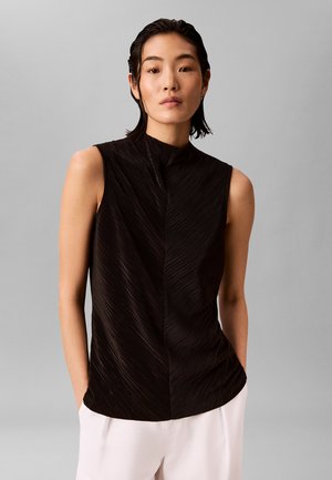 Joven mujer con cabello corto y oscuro que lleva una blusa negra sin mangas con textura y pantalones blancos, de pie frente a un fondo gris liso.