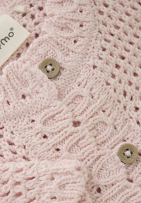 Ljusrosa stickad plagg med ett texturerat spetsmönster med små hål, fäst med en rund, ljusfärgad knapp.