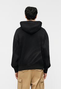 Άτομο που φαίνεται από πίσω, φορώντας μαύρο φούτε ρ με κουκούλα κατεβασμένη και μπεζ παντελόνι cargo, πάνω σε απλό φωτεινό φόντο.