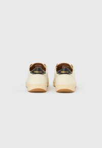 Ténis de couro creme com forma arredondada, solas de borracha cor de castanho-claro e um detalhe preto no calcanhar com o logótipo "Blauer".