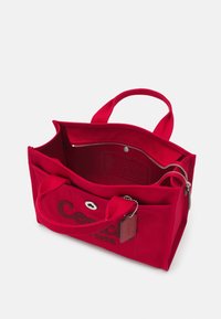Sac fourre-tout rectangulaire rouge avec deux anses, poche avant avec bouton, compartiment principal avec fermeture éclair et petite étiquette attachée.