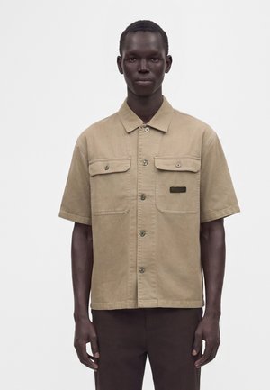 Mannelijk model draagt een beige kort mouw hemd met knopen en twee borstzakken, en een donkere broek, staand tegen een effen achtergrond.
